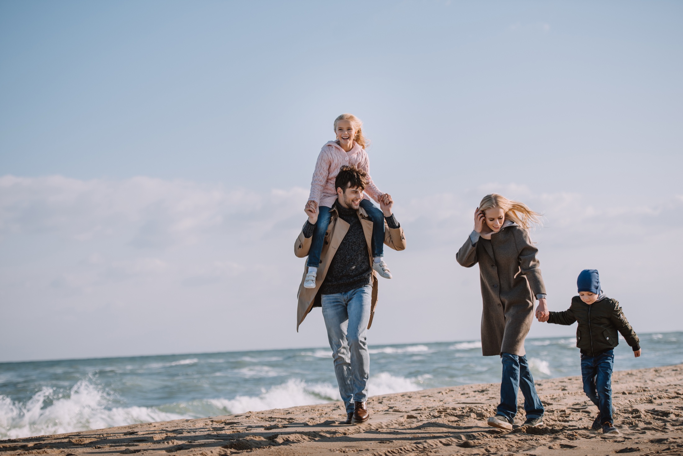 A family walking together along the beach on a bright, breezy day, the father carrying his daughter on his shoulders.