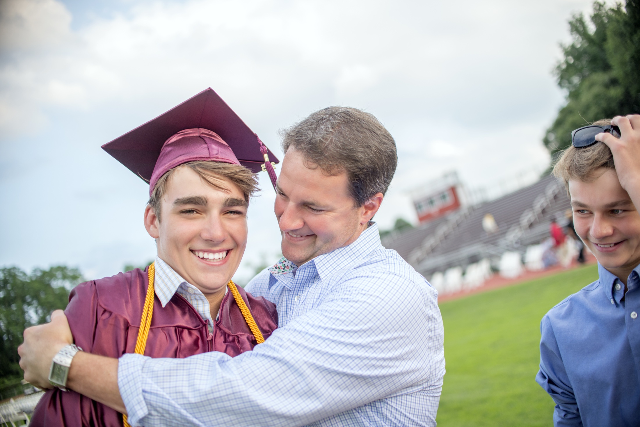 A father embracing his son who is wearing a graduation cap and gown, outdoors at a school stadium.