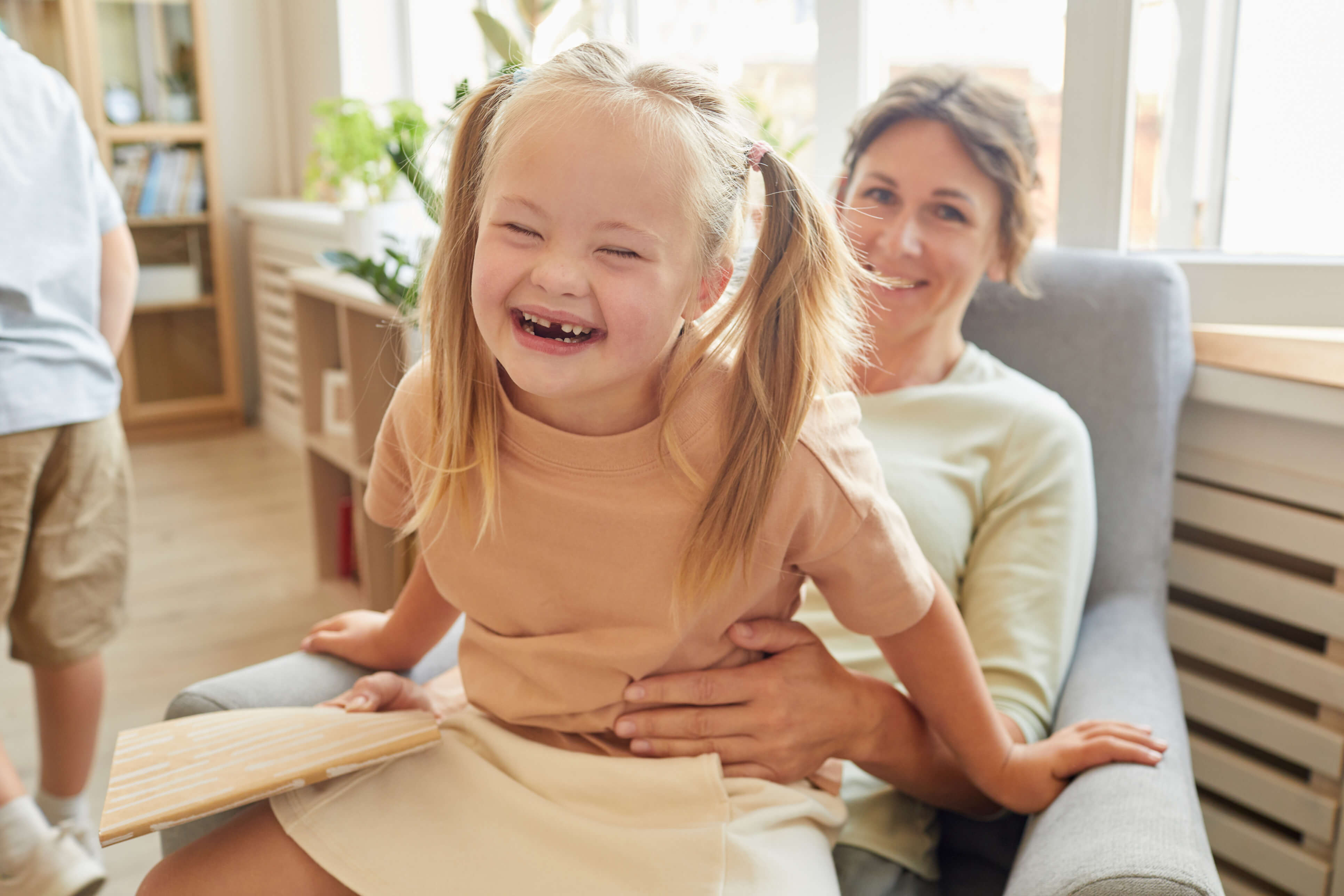 A young girl laughing on a couch with her mother gently holding her, warmly lit by afternoon window light