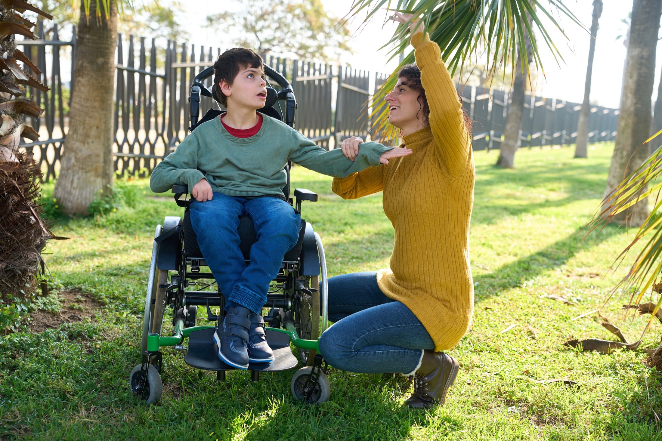 A woman kneeling beside a boy who uses a wheelchair, together outdoors in a park.
