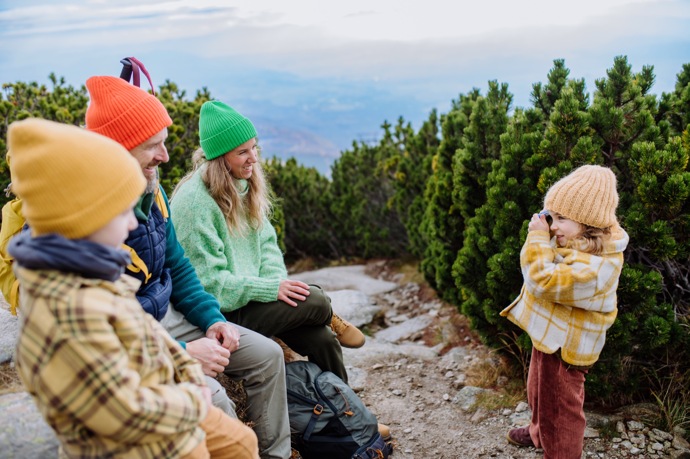 A family resting on a mountain trail as a young child takes their photo, with evergreens and a hazy valley beyond.